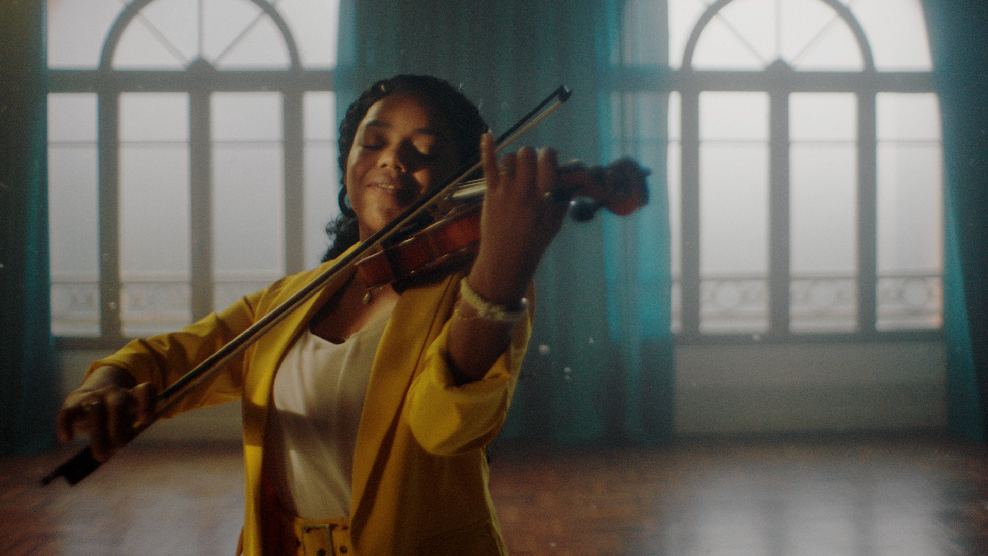 Na imagem, uma mulher vestida de blusa amarela toca violino com leve sorriso no rosto.